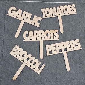 May include: Five wooden plant markers with the words "Garlic", "Tomatoes", "Carrots", "Peppers", and "Broccoli" written on them. The markers are all in a light brown color and have a simple, rustic design.