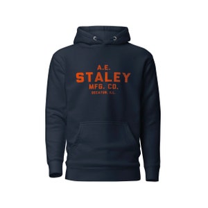 May include: A navy blue hoodie with a white drawstring and a pocket on the front. The hoodie has an orange print that says "A.E. STALEY MFG. CO. DECATUR, ILL."