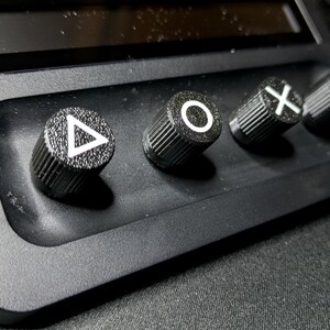 May include: Close-up of four black control knobs with white symbols: triangle, circle, X, and square. The knobs are mounted on a black surface.