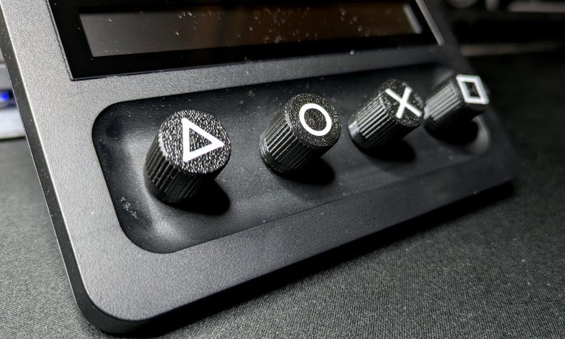 May include: Close-up of four black control knobs with white symbols: triangle, circle, X, and square. The knobs are mounted on a black surface.