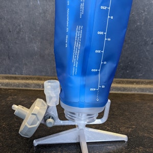 Op de afbeelding: Een blauwe waterhydratatieblaas met een doorzichtige tuit en een witte dop. De blaas is bevestigd aan een zilveren standaard met drie poten. De blaas heeft markeringen voor 200, 250, 300, 350, 400 en 450 milliliter.