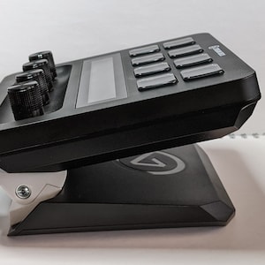 Puede incluir: Un controlador de m&uacute;sica negro con un soporte blanco. El controlador tiene cuatro botones, una peque&ntilde;a pantalla y una cuadr&iacute;cula de botones. El soporte tiene un logotipo en la parte inferior.