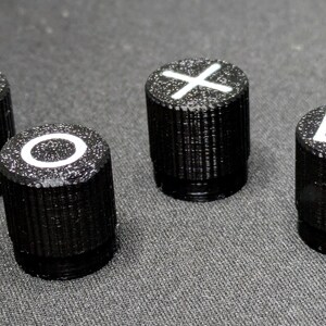 May include: Four black plastic buttons with white symbols. The symbols are a triangle, a circle, an X, and a square.