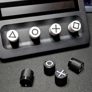 May include: A set of six black and white buttons with different symbols. The buttons are round and have a raised edge. The symbols are a triangle, a circle, a cross, a square, a circle with a dot in the centre, and a cross with a dot in the centre.