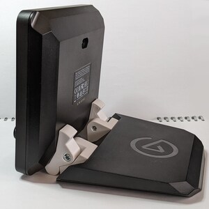 Puede incluir: Un soporte negro con una bisagra blanca y un logotipo en la parte delantera. El soporte est&aacute; dise&ntilde;ado para sostener una tableta o un tel&eacute;fono.