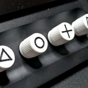 May include: Four white round buttons with black symbols on a black surface. The symbols are a triangle, a circle, a plus sign, and a square.