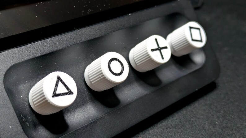 May include: Four white round buttons with black symbols on a black surface. The symbols are a triangle, a circle, a plus sign, and a square.