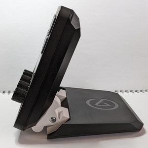 Puede incluir: Una interfaz de audio negra con un soporte plegable. La interfaz tiene cuatro botones giratorios y varios botones. El soporte tiene una bisagra blanca y un logotipo en la parte inferior.