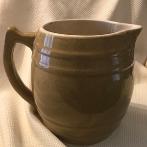 May include: A brown ceramic pitcher with a wide mouth and a handle. The pitcher has a glazed finish and is shaped like a barrel.