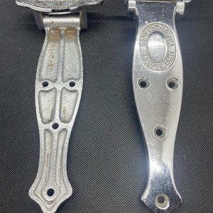 May include: Two vintage silver-toned metal hinges. One hinge has a textured top and the other has an oval emblem with the text "BALTIMORE OTTENHEIMER BROS". Both hinges have a decorative, elongated design with multiple screw holes.