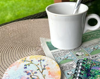 Puede incluir: Una taza de café de cerámica blanca con una cuchara plateada en su interior se encuentra sobre una mesa junto a un posavasos floral y una tira cómica de periódico. El posavasos presenta una pintura de una rama con flores azules sobre un fondo pastel.