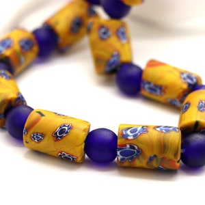 Könnte beinhalten: Nahaufnahme einer Halskette aus gelben und blauen Glasperlen. Die Perlen sind zylindrisch und haben ein wirbelndes Muster. Die Halskette liegt auf einem weißen Hintergrund.