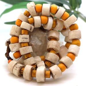 Peut inclure: Un collier composé de perles rectangulaires crème alternant avec des perles rondes orange. Le collier est enfilé sur un cordon marron foncé. Les perles ont un aspect rustique et naturel, sur fond neutre.