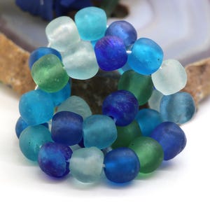 Può includere: Una collezione di perline arrotondate con finitura opaca in varie tonalità di blu, verde acqua e verde. Le perline hanno un aspetto satinato simile al vetro marino con forme organiche irregolari, raggruppate in un insieme di toni freddi.