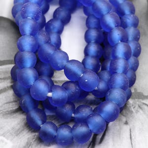 Può includere: Primo piano di fili di perline rotonde, opache, blu cobalto. Le perline sono infilate su uno spago bianco. Lo sfondo è un'immagine sfocata in bianco e nero.