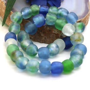 Può includere: Una collana di perline in vetro smerigliato e arrotondato in varie tonalità di blu, verde e bianco traslucido. Le perline hanno una consistenza opaca e usurata, infilate su un sottile cordino bianco, creando un aspetto organico e multicolore.