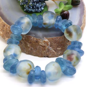 Könnte beinhalten: Ein elastisches Armband bestehend aus abwechselnd mattierten hellblauen Glasperlen und kleineren, durchscheinenden blauen Glaszwischenperlen. Die größeren Perlen haben eine weiche, matte Oberfläche mit subtilen, wolkigen weißen und braunen Einschlüssen.