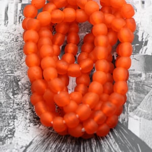 Könnte beinhalten: Nahaufnahme einer Kette aus orangefarbenen Perlen. Die ovalen Perlen haben eine matte Oberfläche. Die Farbe ist ein leuchtendes Orange. Die Perlen sind aufgefädelt, möglicherweise für eine Halskette oder anderen Schmuck. Der Hintergrund ist ein monochromer Muster.