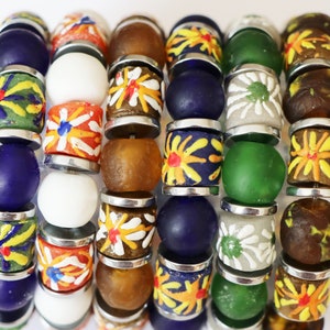 Puede incluir: Una colección de cuentas de vidrio coloridas con varios diseños. Las cuentas incluyen colores sólidos como azul, blanco y verde, así como cuentas con patrones florales en amarillo, naranja y blanco. Cada cuenta está separada por un espaciador de metal plateado.
