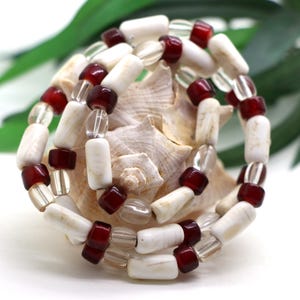 May include: A beaded bracelet featuring a repeating pattern of cylindrical white beads, deep red square beads, and clear translucent beads. The beads are strung together in a circular arrangement, showcasing a contrast between the opaque white, glossy red, and transparent glass elements throughout the piece.