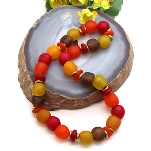 Pode incluir: Um colar de contas com elementos mate e arredondados em tons de vermelho, laranja, amarelo e castanho. As contas são intercaladas com pequenos espaçadores planos de cor dourada metálica, criando um padrão repetitivo de cores quentes.