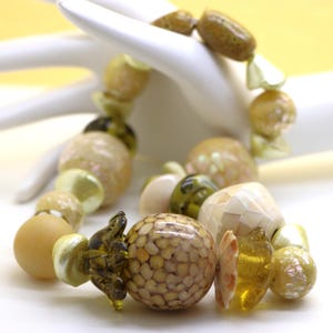 Peut inclure: Un bracelet de perles composé de perles rondes et irrégulières dans des tons crème, beige, vert olive et ambre. Les perles incluent des sphères mates lisses, des pièces en verre translucide et des éléments texturés avec des motifs nacrés.