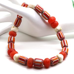 May include: A necklace featuring a variety of beads in red, white, and orange. The necklace includes cylindrical beads with striped patterns, round red beads, and off-white beads. The necklace is displayed on a white surface.