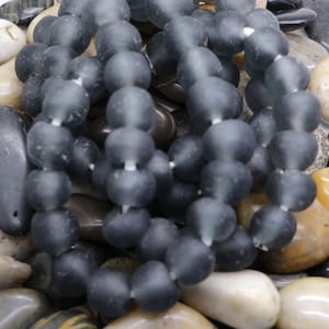 Op de afbeelding: Close-up van strengen matte grijze glazen kralen. De kralen zijn rond en geregen op een wit koord. De kralen liggen op een bed van gladde, veelkleurige stenen. De kralen hebben een matte uitstraling.