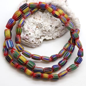 Può includere: Fili di perline colorate a forma di botte. Le perline presentano un vivace mix di strisce rosse, gialle, verdi, blu e nere. Le perline sono infilate insieme, creando una collana o un braccialetto. Lo sfondo include una conchiglia bianca.