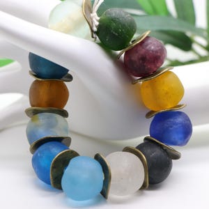 Könnte beinhalten: Ein farbenfrohes Armband mit einer Vielzahl von Glasperlen in Blau-, Gelb-, Grün- und Lilatönen. Die Perlen sind durch kleine, runde, bronzefarbene Metallakzente getrennt. Das Armband ist aufgefädelt und schafft ein lebendiges und auffälliges Accessoire.