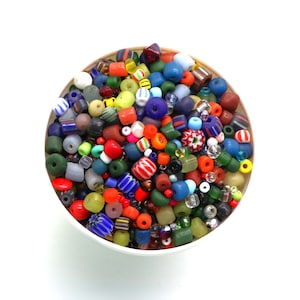 Op de afbeelding: Een ronde bak gevuld met een grote variëteit aan losse kralen in diverse vormen, maten en kleuren, waaronder rood, blauw, groen, geel, wit en zwart, met effen, doorschijnende, gestreepte en gedessineerde glazen ontwerpen.
