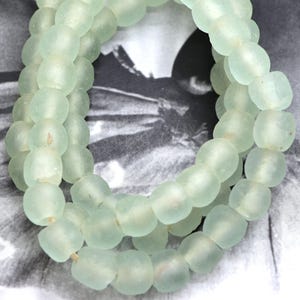 May include: Strands of pale green, rounded glass beads. The beads have a slightly frosted appearance and are strung together, creating a necklace or bracelet. The background is a blurred black and white image.