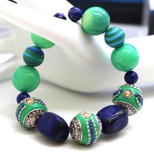 Puede incluir: Una pulsera de cuentas colorida con cuentas verdes, azules y plateadas. La pulsera incluye cuentas verdes grandes y redondas, cuentas azules y verdes a rayas y cuentas ovaladas azul oscuro. Algunas cuentas tienen detalles plateados ornamentados y pequeños detalles azules.