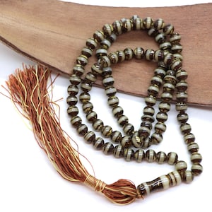 Può includere: Una collana di perline con perline a righe marroni e bianche. La collana ha un nappa fatta di fili marroni e dorati. Le perline sono infilate insieme e la collana è esposta su una superficie di legno.