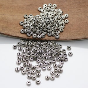 Può includere: Un grande mucchio di perline di metallo color argento con un bordo decorativo a festoni. Le perline sono rotonde con un foro al centro.
