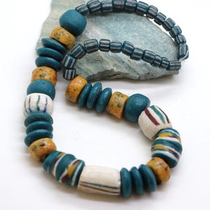Puede incluir: Un collar con una variedad de cuentas coloridas. Las cuentas son en tonos turquesa, amarillo y blanco, con patrones rayados y moteados. El collar se exhibe sobre una piedra verde claro.