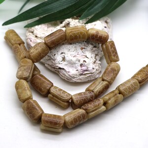 May include: A strand of rectangular beads in a light brown, tan color. Each bead features a carved, grid-like pattern on its surface that resembles a turtle shell texture. The beads are strung together in a loose, curved arrangement.
