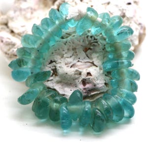 Puede incluir: Un hilo circular de cuentas de vidrio translúcido de color azul claro con formas irregulares y aplanadas. Las cuentas están ensartadas en un cordón blanco fino, mostrando tonos variados de aguamarina y cerceta con una textura suave y pulida.