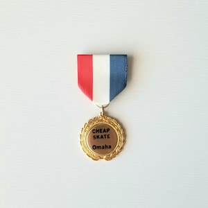 May include: A gold-colored medal with a laurel wreath design and the words "CHEAP SKATE Omaha" on a brown background. The medal hangs from a red, white, and blue ribbon.