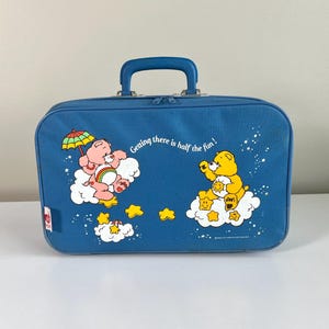 May include: A blue rectangular suitcase with a top handle and zipper closure. The front features a graphic of two Care Bears on clouds, one pink and one yellow, with the text "Getting there is half the fun!" surrounded by small yellow stars.