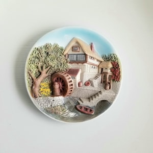 May include: A ceramic wall plate with a 3D design of a cottage with a thatched roof, a water wheel, and a stone bridge. The plate is painted in shades of brown, green, and blue.