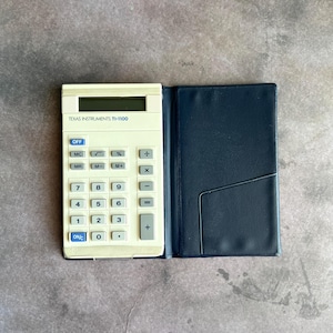 May include: A vintage Texas Instruments TI-1100 calculator with a cream-colored body and a dark blue leatherette case. The calculator has a digital display and a full set of number and function keys. The case is open, revealing the calculator.