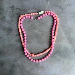 May include: Two pink necklaces with varying bead sizes and shapes. The top necklace features small, round beads, while the bottom necklace has larger, round beads. Both necklaces have a mix of light and dark pink beads. The necklaces are connected with a silver clasp.