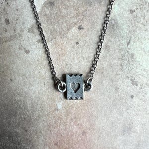 May include: A silver-toned necklace featuring a small, rectangular pendant with a heart cutout. The pendant has a decorative, notched edge. The chain is delicate and links to the pendant with small loops.