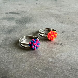May include: Two adjustable silver rings, each with a colorful, textured pom-pom design. One ring features a pom-pom in shades of pink and blue, while the other has a pom-pom in orange and yellow. The rings are set against a gray background.