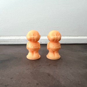 May include: Two small, peach-colored figurines of babies wearing diapers. The figurines are standing on a dark brown surface.