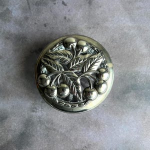 May include: A silver-toned, round decorative box with a detailed relief of cherries and leaves. The lid features a raised design of cherries and foliage, creating a three-dimensional effect. The box has a shiny, reflective surface.