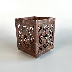 May include: A square metal candle holder featuring a dark, weathered, rust-colored finish. Each side displays a symmetrical, intricate cut-out pattern resembling a stylized floral or quatrefoil design, creating an openwork effect throughout.