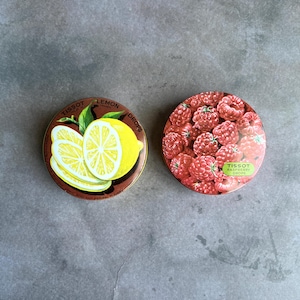 May include: Two round tins of Tissot drops. One tin features a lemon design with the text "Lemon Drops". The other tin is filled with raspberries and the text "Raspberry Drops". Both tins have a brown background.