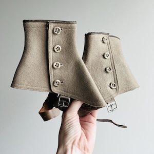 May include: A pair of beige felt spats featuring four round buttons on the side, dark brown leather trim along the top edges, and adjustable brown leather straps with silver-toned metal buckles at the bottom for securing around the footwear.
